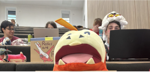 Pokémon plushie on a table with people with Pokémon hats and signs in the background
